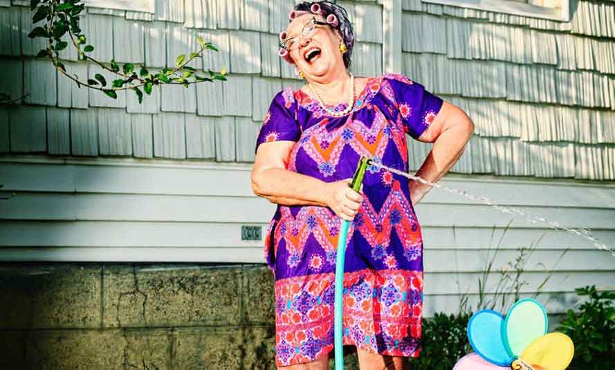 Senior woman with vibrant purple and pink dress and hair in rollers, laughing while watering her garden