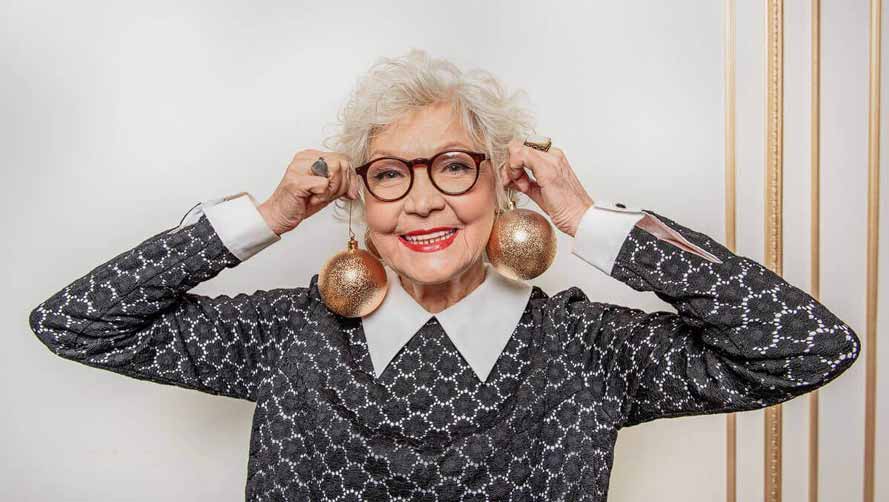 Smiling senior woman with black glasses, holding two gold Christmas baubles up as earrings