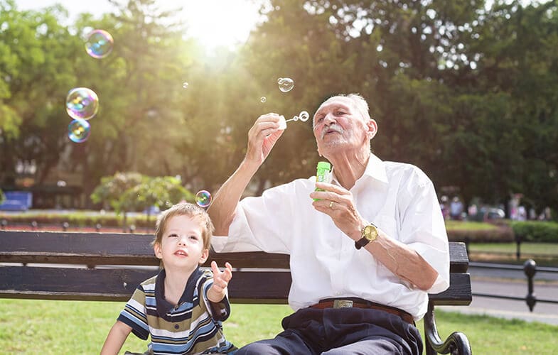 Older man sitting on a park bench blowing bubbles with his grandson