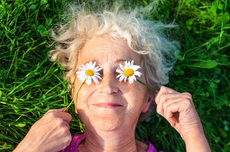 Smiling senior woman lying in the grass and holding daisies over her eyes