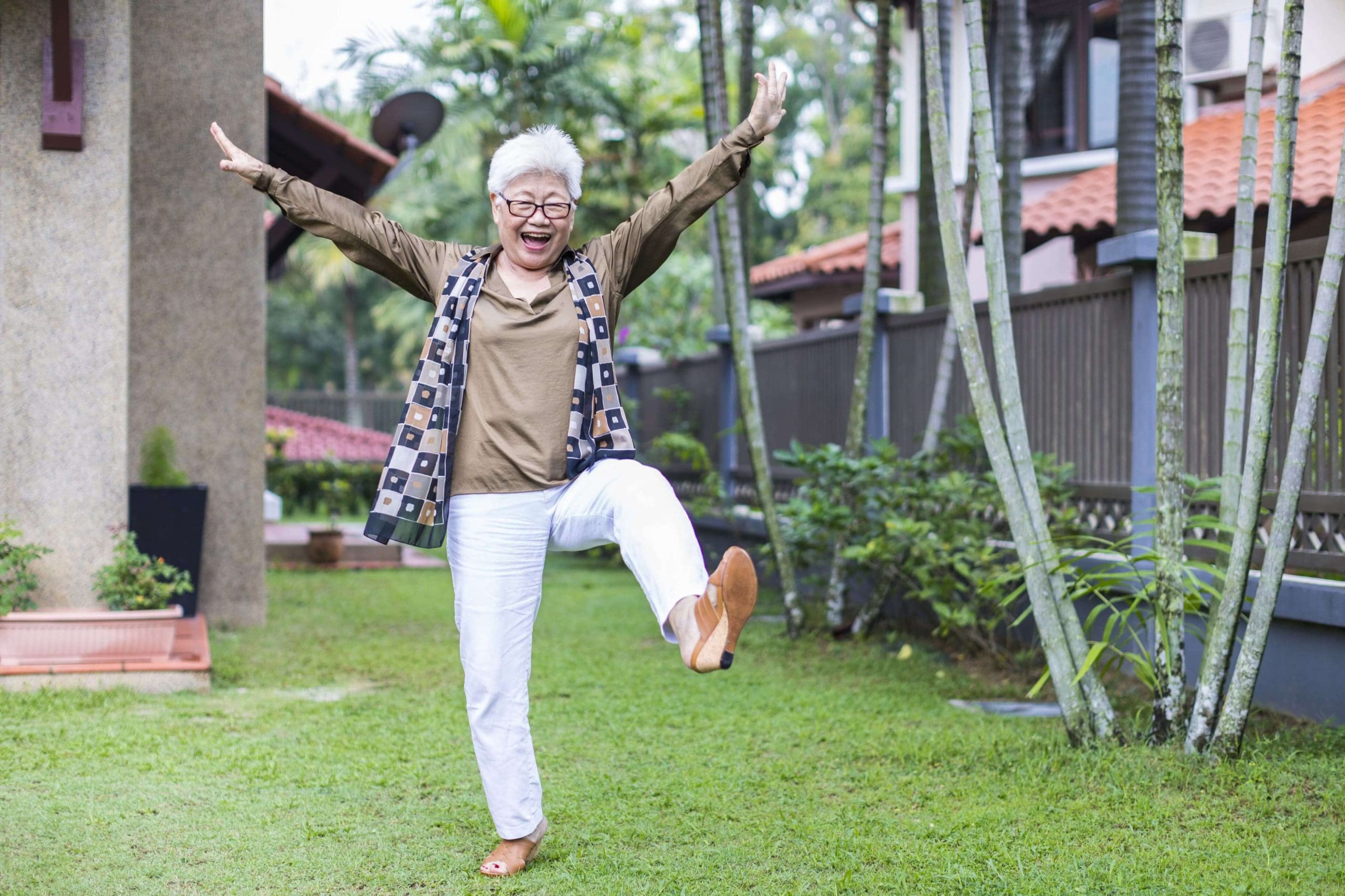 Older Asian woman dancing in backyard, happy with arms raised