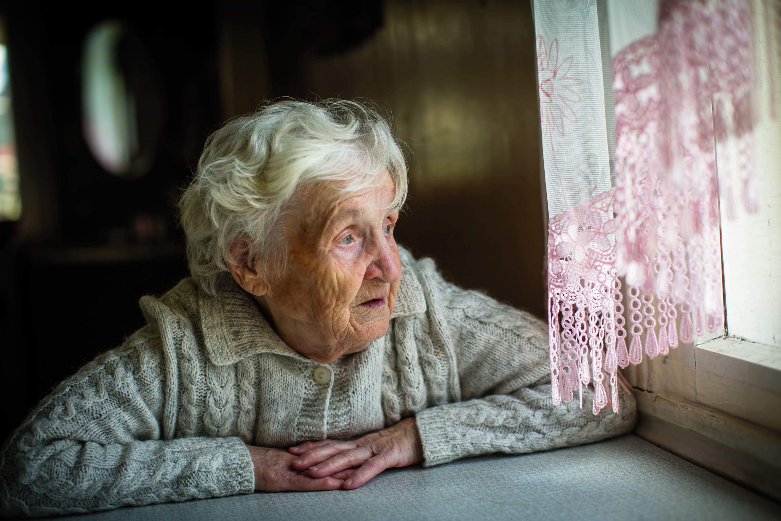 Elderly woman sits at a table staring longingly out the window