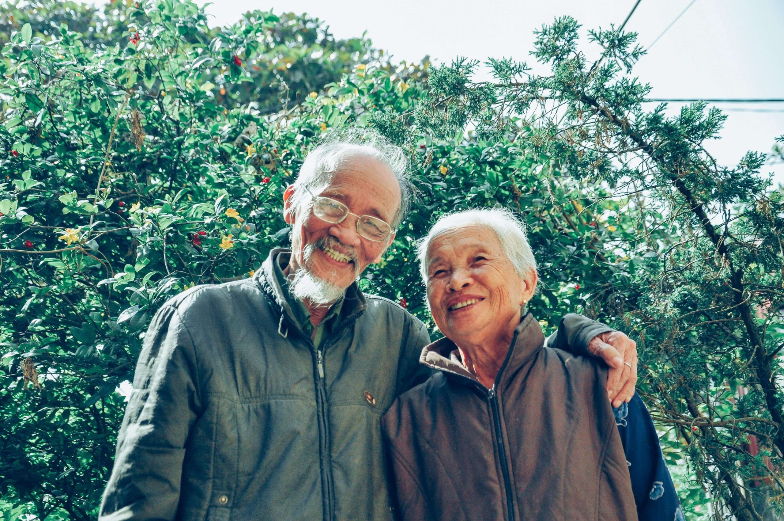 Smiling elderly couple standing in front of a tall hedge of trees