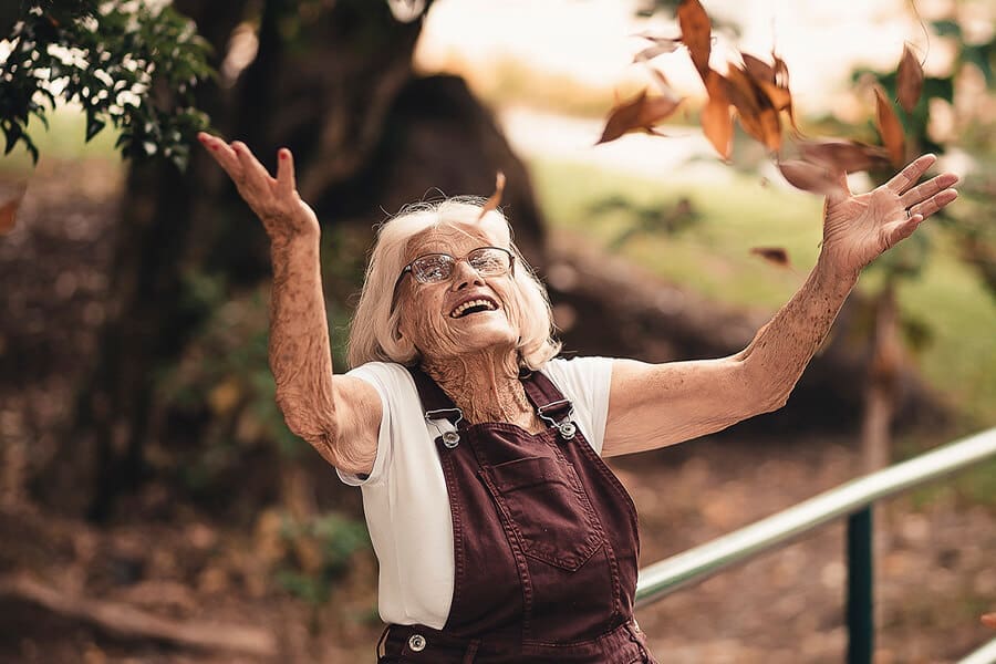 Senior lady in brown overalls throwing autumn leaves into the air