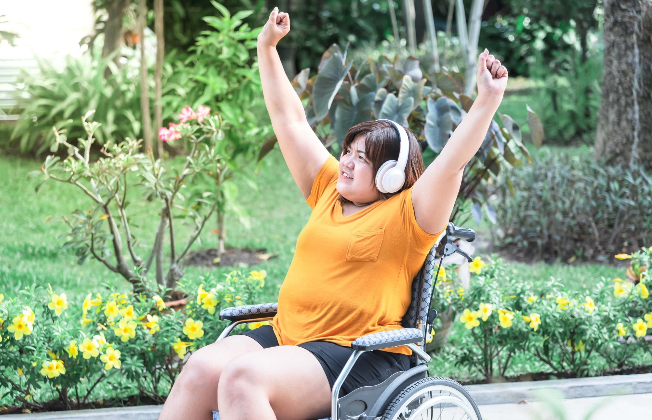 Young woman in a wheelchair with headphones on and arms raised in the air in triumph