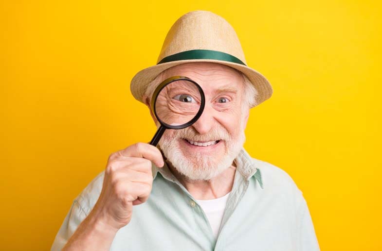 An older man holding a magnifying glass up to his eye and smiling